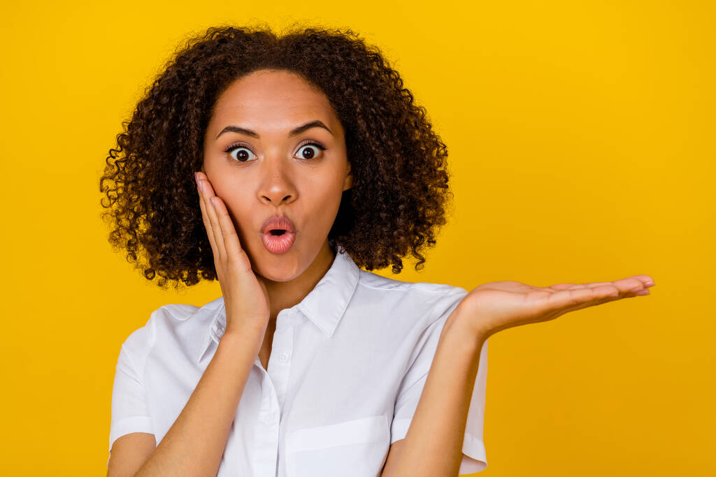 Photo of astonished impressed pretty girl with curly hairstyle arm cheekbone showing empty space isolated on yellow color background. - Foto, Bild