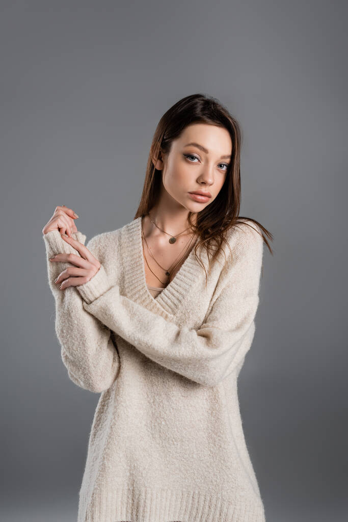 young woman in cozy sweater and golden necklaces looking at camera isolated on grey - Foto, Imagem