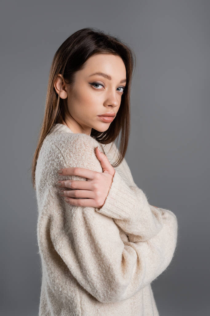 brunette woman in soft sweater hugging herself and looking at camera isolated on grey - Foto, Bild