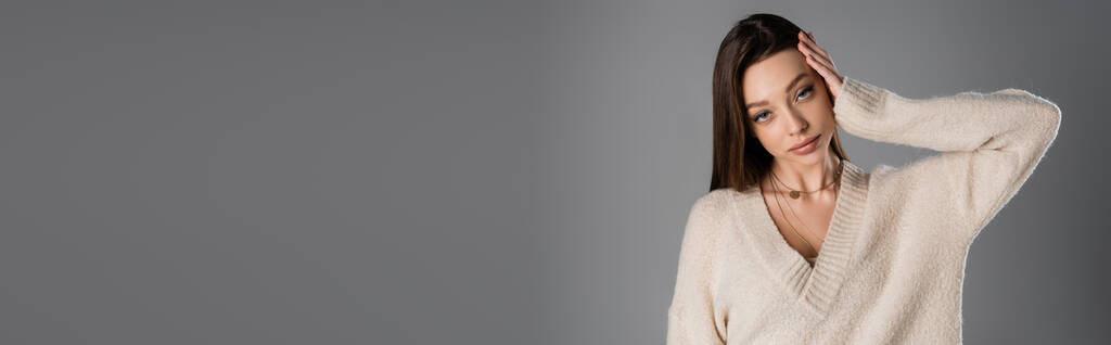brunette woman in cozy sweater touching head and looking at camera isolated on grey, banner - Foto, Imagem