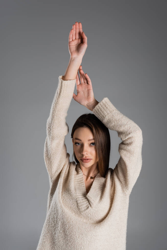 sensual woman in warm sweater standing with raised hands and looking at camera isolated on grey - Fotó, kép