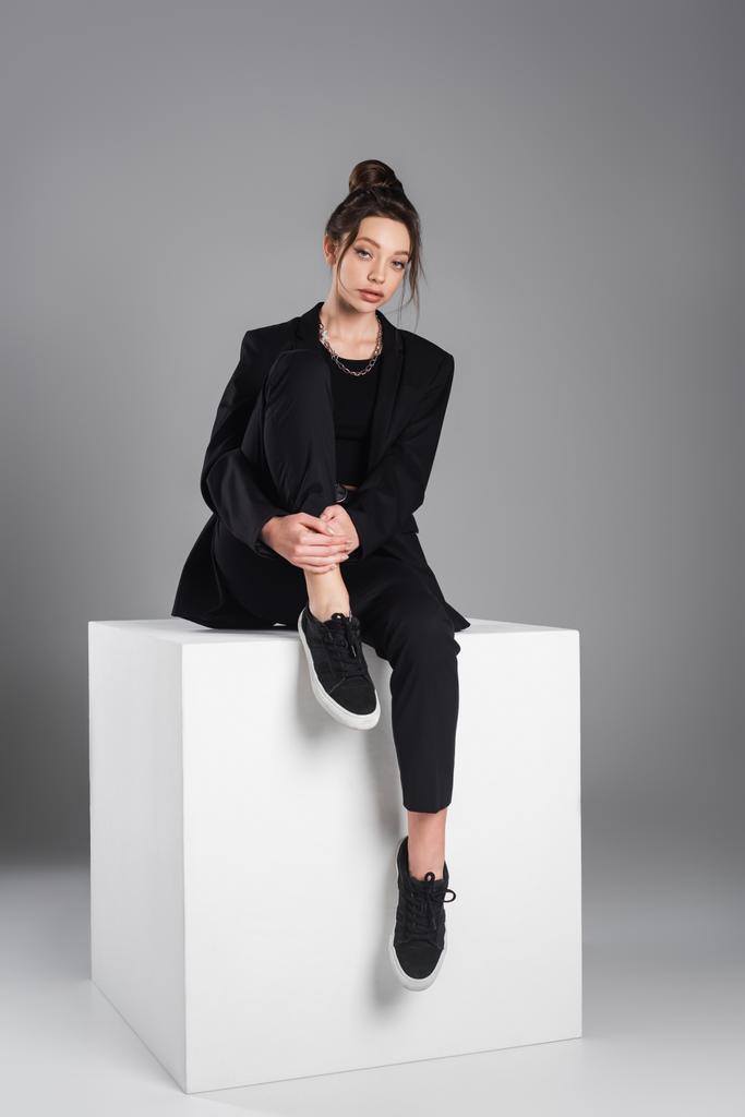 full length of woman in black suit and sneakers sitting on white cube on grey background - Foto, afbeelding