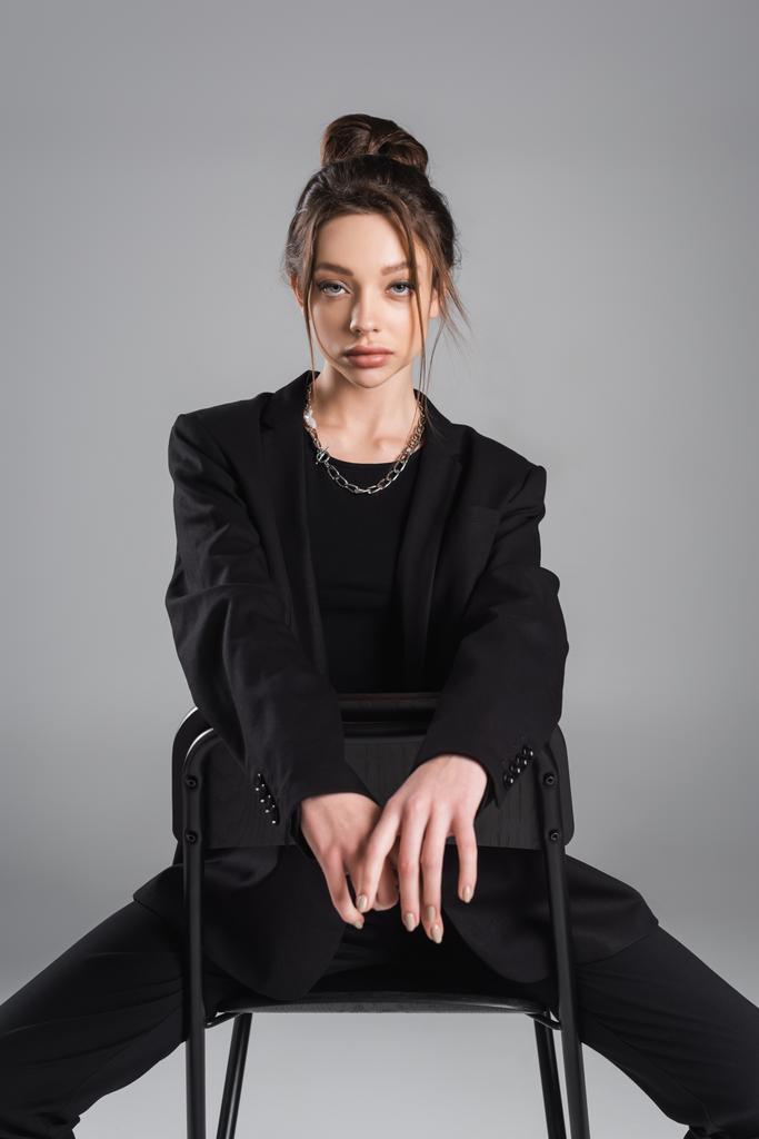 brunette woman in black clothes sitting on chair and looking at camera isolated on grey - Zdjęcie, obraz