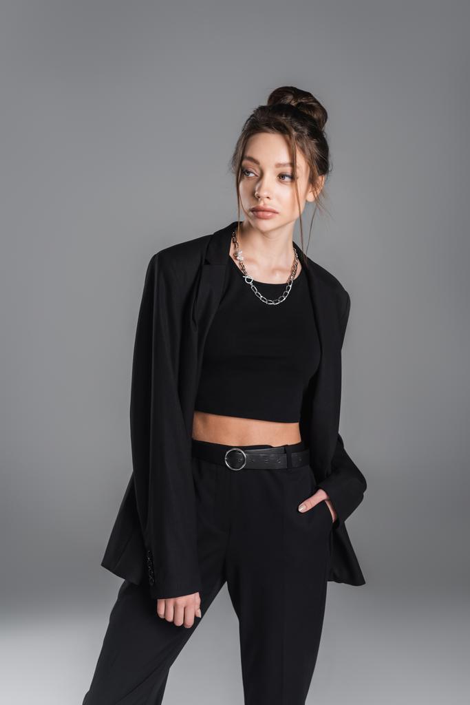 young and stylish woman in black clothing holding hand in pocket and looking away isolated on grey - Foto, imagen