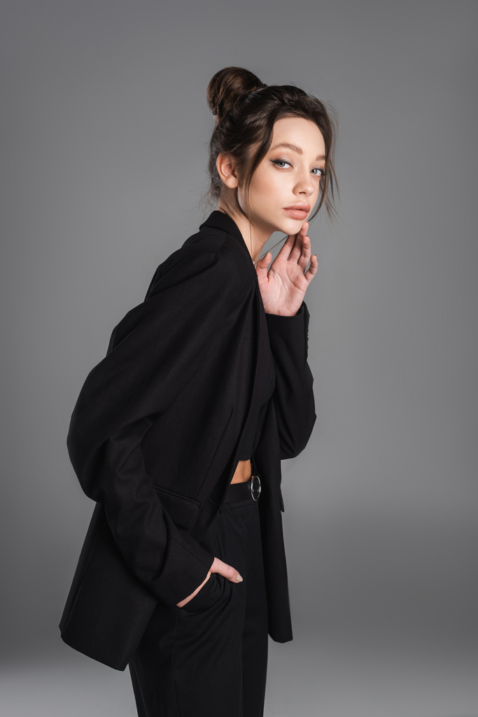 young woman in black clothes standing with hand in pocket and touching face isolated on grey - Foto, immagini