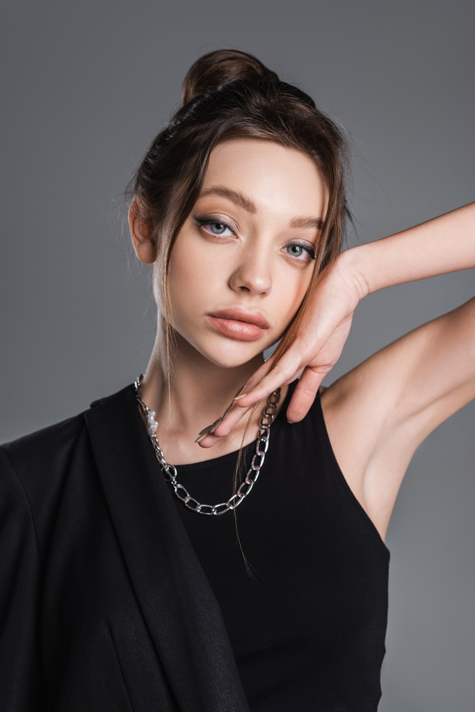 young woman with makeup and silver necklace posing with hand near face isolated on grey - Foto, imagen