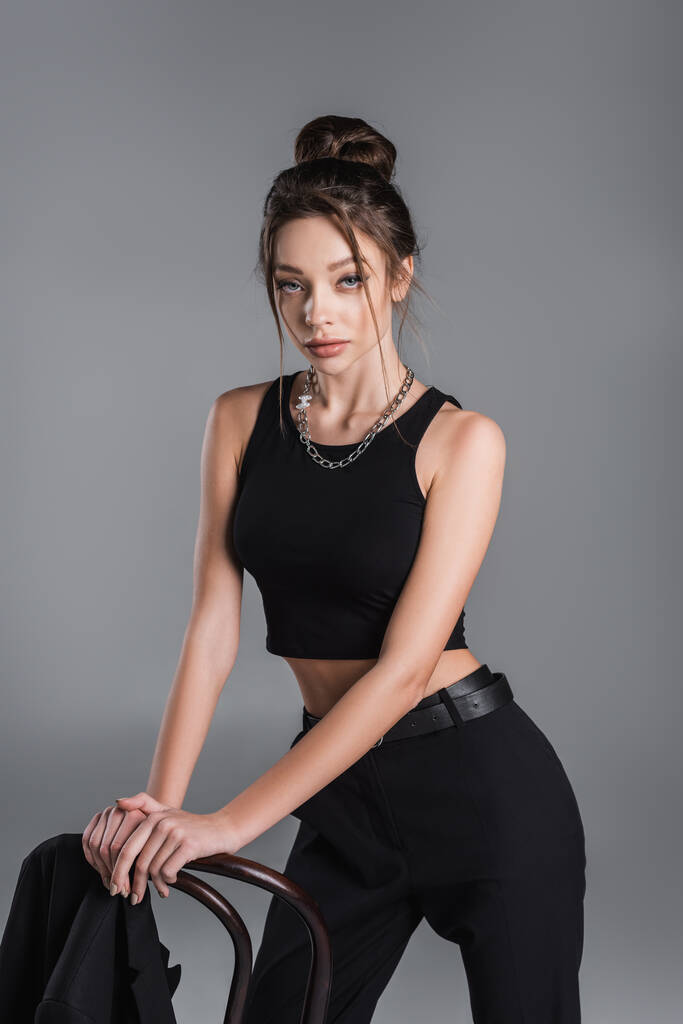 trendy woman in black crop top and silver necklace posing near chair isolated on grey - Valokuva, kuva
