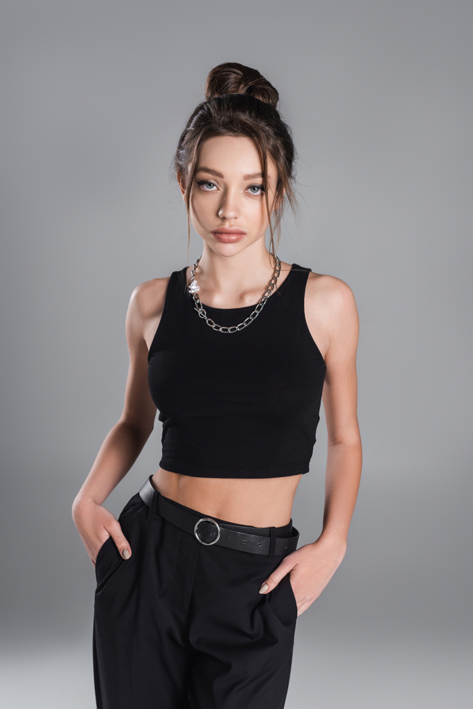 young woman in black crop top posing with hands in pockets of trousers isolated on grey - Zdjęcie, obraz