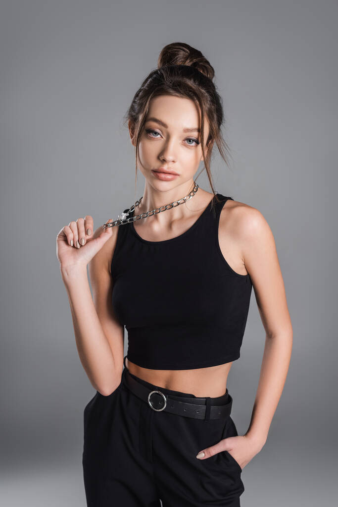 fashionable woman in black crop top touching metal necklace and looking at camera isolated on grey - Photo, Image