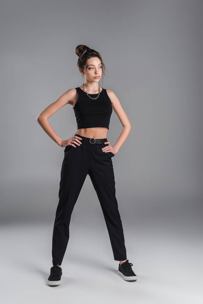 full length of young woman in black crop top and trousers posing with hands on hips on grey background - 写真・画像