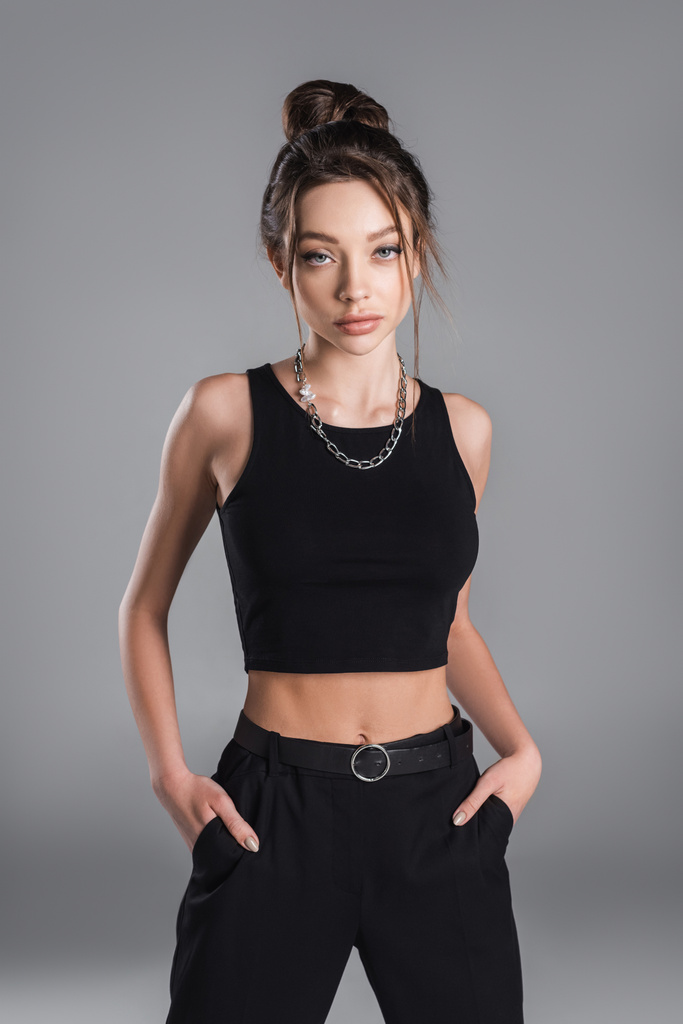 brunette woman in black crop top posing with hands in pockets isolated on grey - Foto, Bild