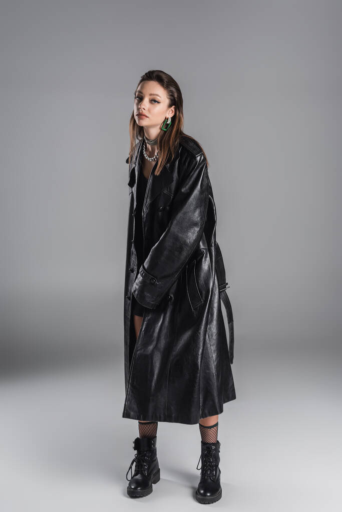 full length of trendy woman in black leather coat on grey background - 写真・画像