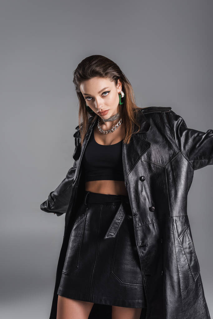 young brunette woman in trendy leather outfit looking at camera while posing isolated on grey - Foto, Bild