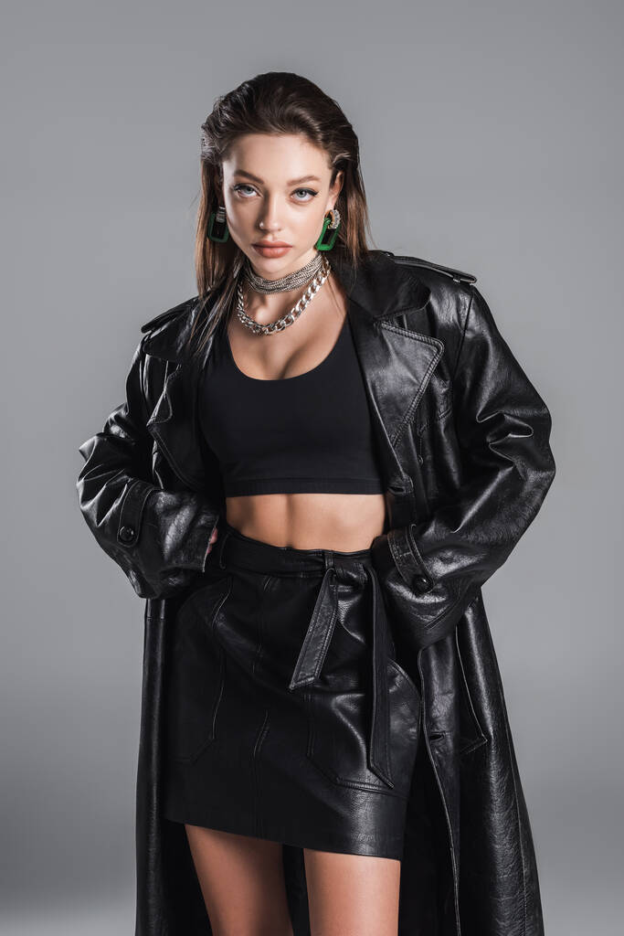 brunette woman in black crop top and leather jacket standing with hands in pockets isolated on grey - Foto, afbeelding