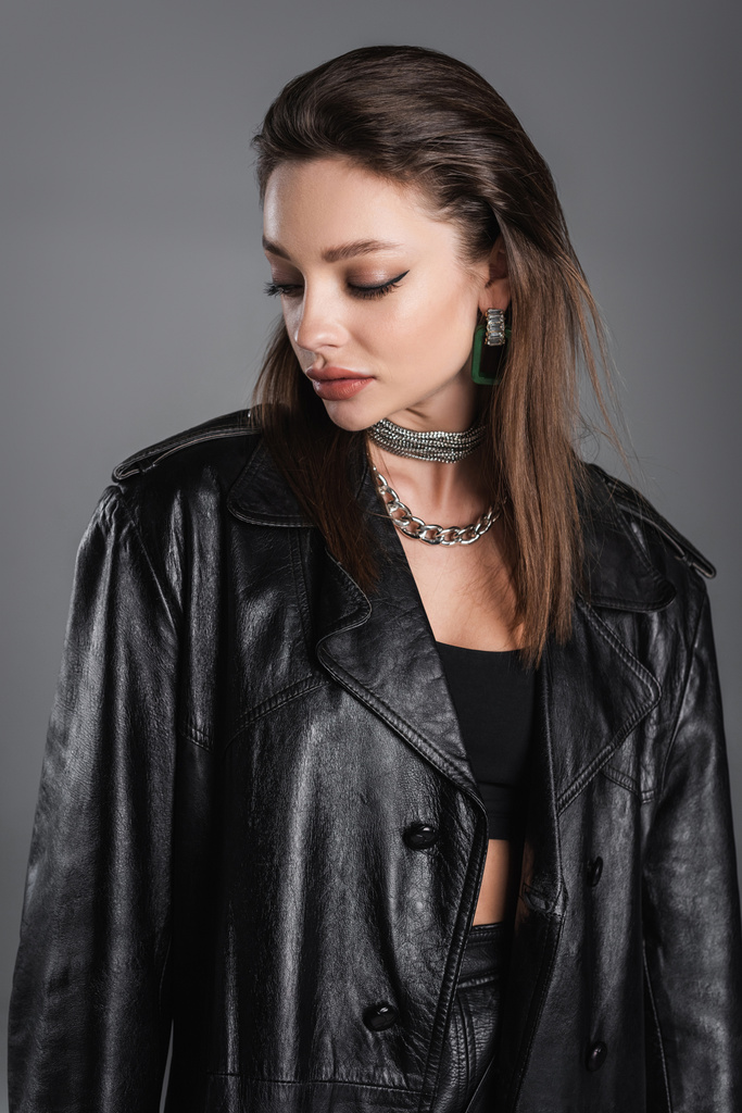 young woman with makeup and silver necklaces posing in black leather coat isolated on grey - Фото, изображение