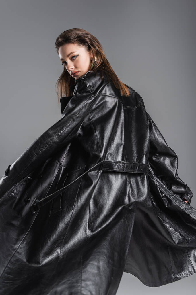young and stylish woman in black leather coat looking at camera while posing isolated on grey - Фото, изображение