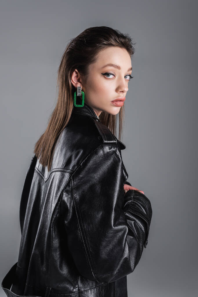 brunette woman with makeup standing in black leather coat and looking at camera isolated on grey - Photo, Image