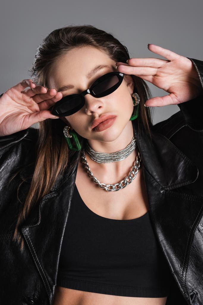 молода жінка в срібних намистах регулює темні і модні сонцезахисні окуляри ізольовані на сірому
 - Фото, зображення