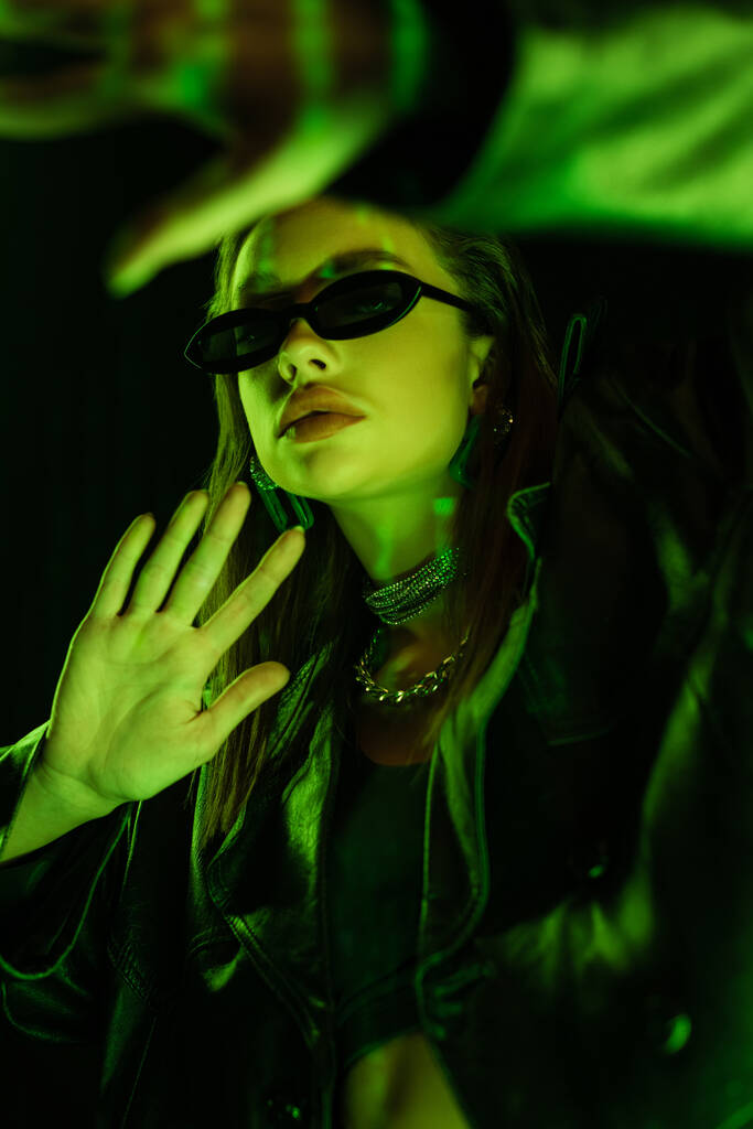 young woman in dark sunglasses gesturing in green neon light isolated on black - Valokuva, kuva