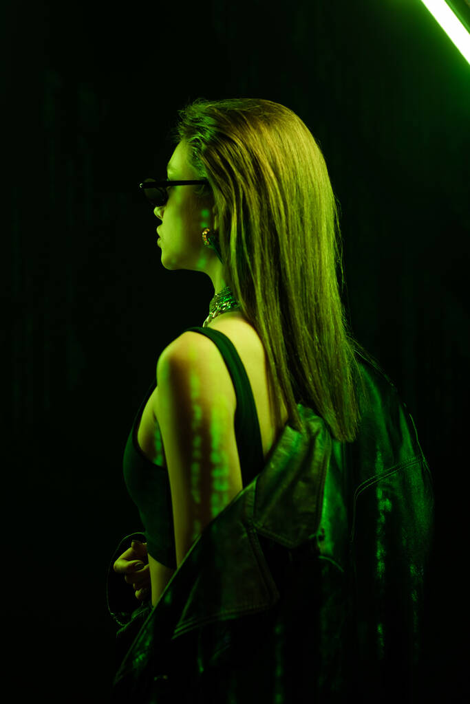 stylish woman in sunglasses posing with leather coat in green neon light on black background - Photo, Image