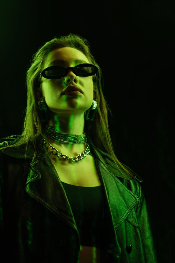 low angle view of young woman in trendy sunglasses and metal necklaces in green neon light isolated on black - Fotó, kép