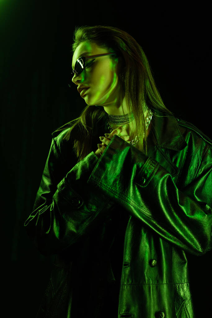 trendy woman in leather coat and dark sunglasses holding hands near chest in green light isolated on black - Foto, Imagen