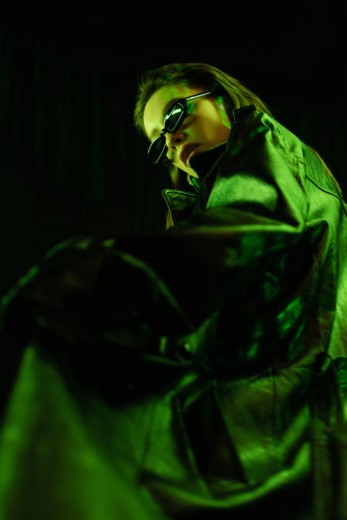low angle view of woman in leather coat and dark sunglasses in green light isolated on black - Fotoğraf, Görsel