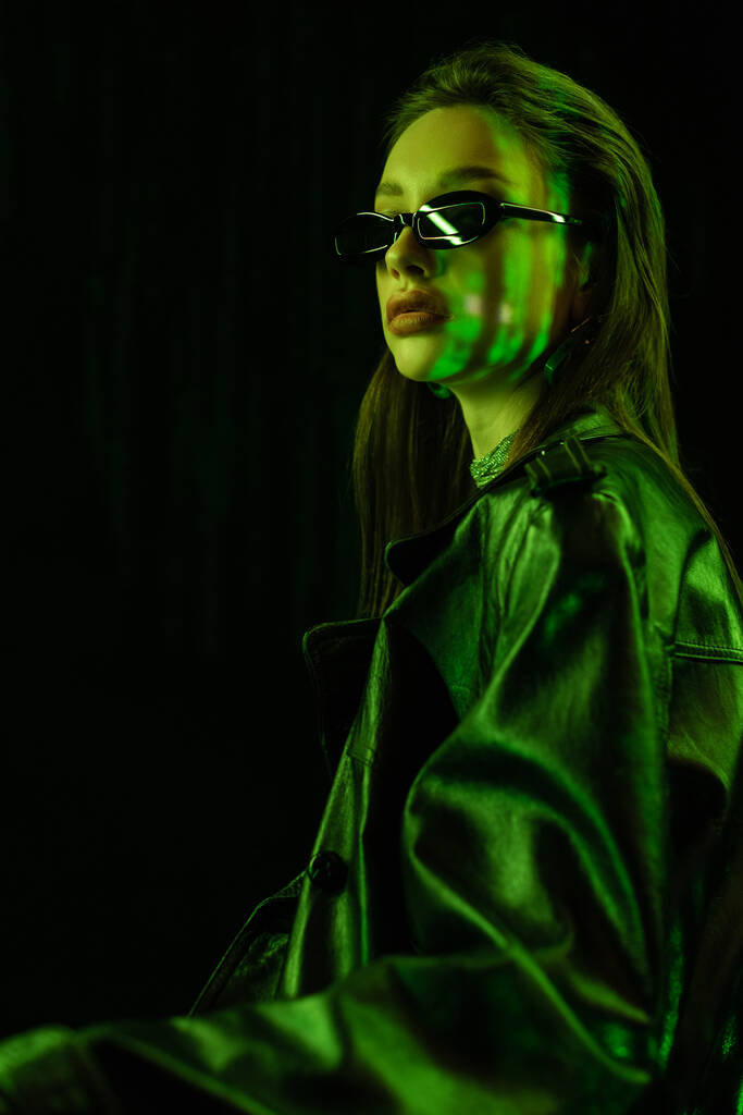 молода брюнетка в шкіряному пальто і сонцезахисні окуляри в зеленому неоновому світлі ізольовані на чорному
 - Фото, зображення