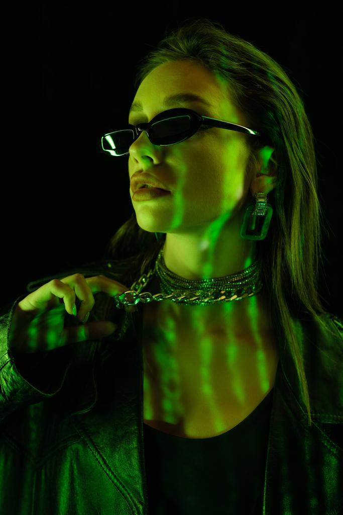 portrait of young woman in dark sunglasses touching silver necklace in green light isolated on black - Foto, afbeelding