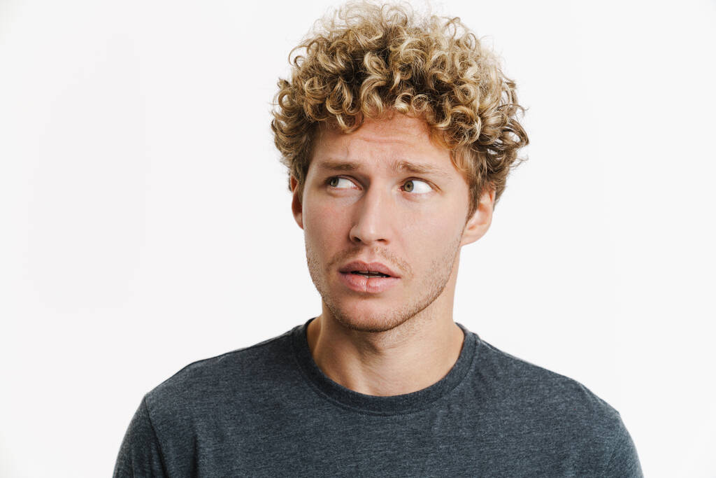 Young puzzled man with curly hair posing and looking aside isolated over white wall - 写真・画像