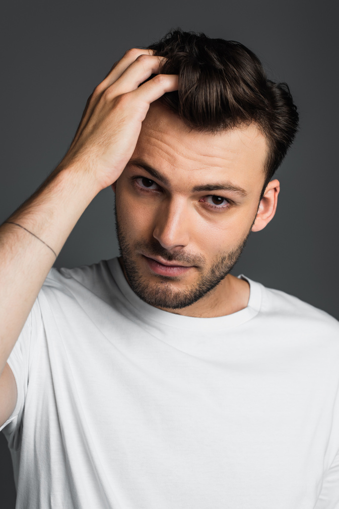 Portrait of brunette man in t-shirt touching hair isolated on grey  - Foto, imagen
