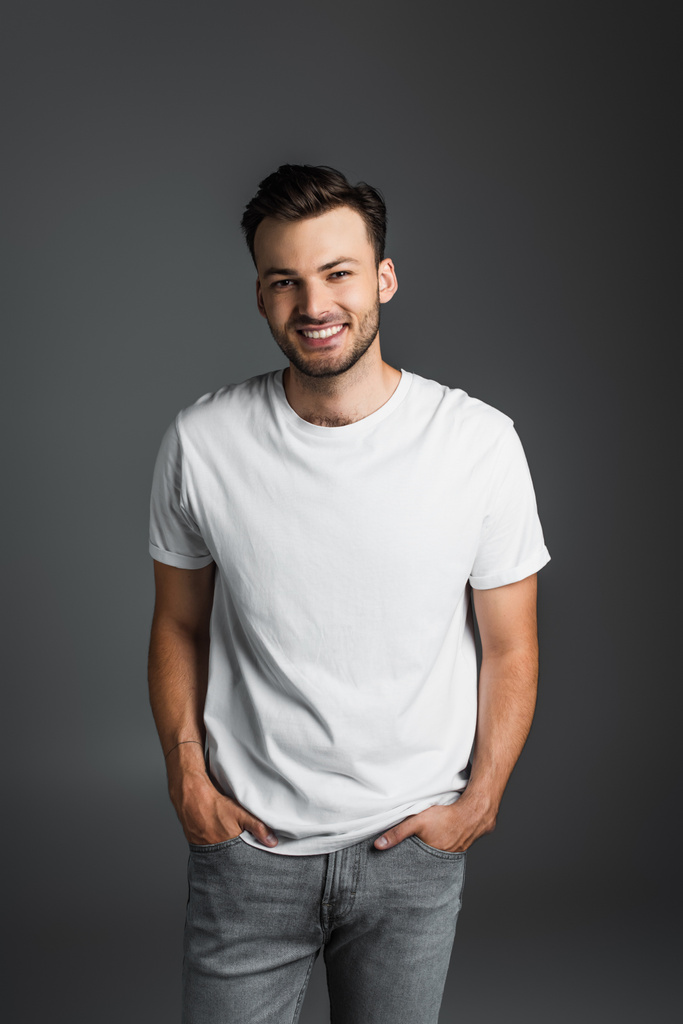 Positive brunette man in white t-shirt and jeans posing isolated on grey  - Zdjęcie, obraz