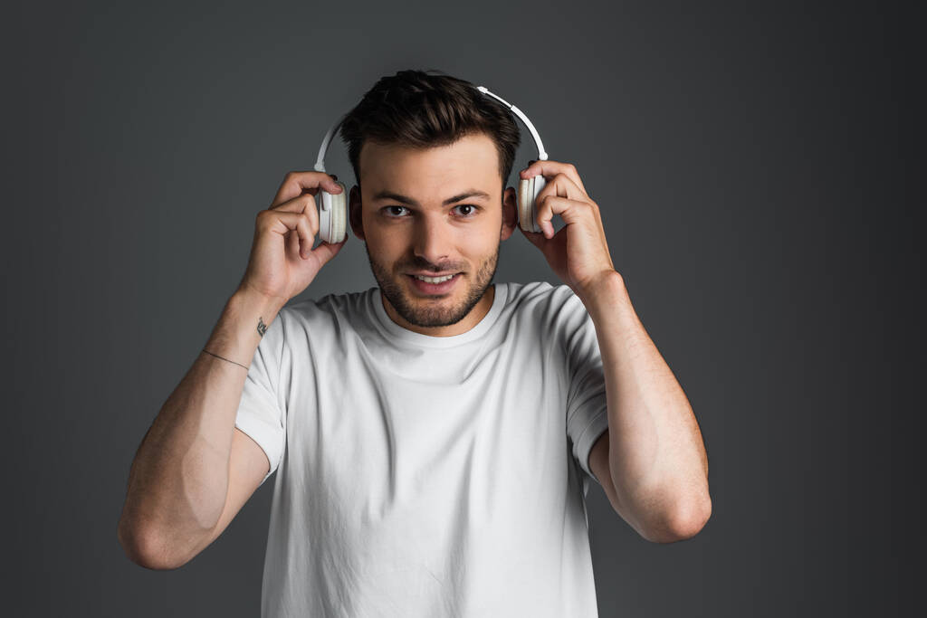 Smiling man in white t-shirt holding headphones near ears isolated on grey  - 写真・画像