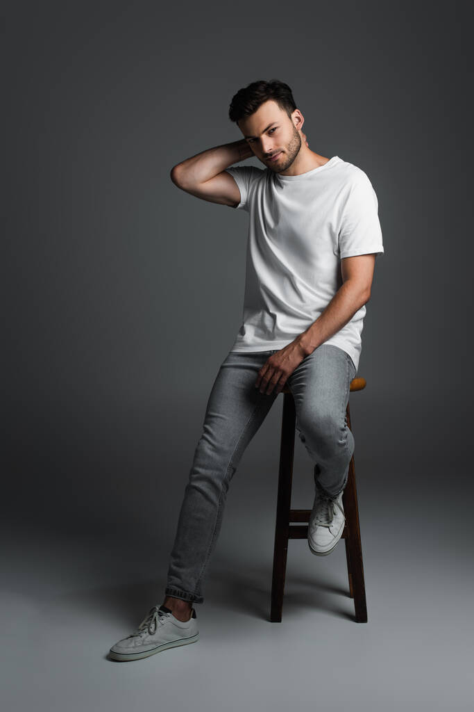 Full length of young man in t-shirt and jeans sitting on chair on grey background - 写真・画像