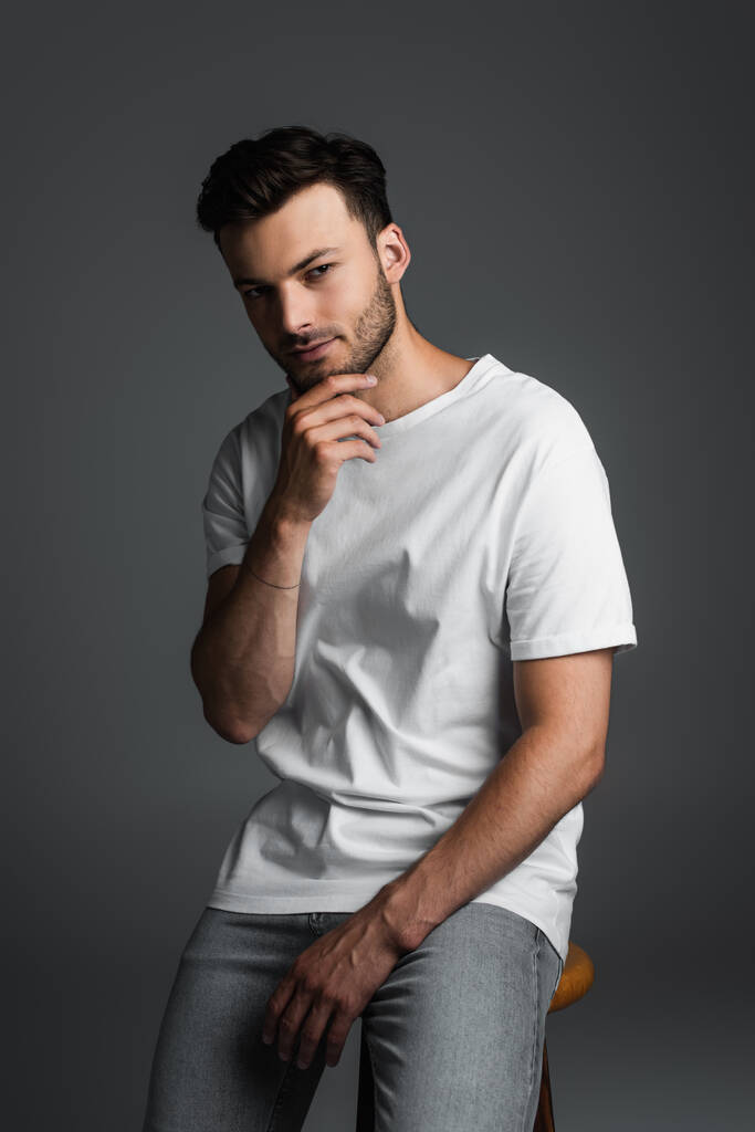 Brunette man in jeans and white t-shirt touching chin while sitting on chair isolated on grey  - Fotografie, Obrázek