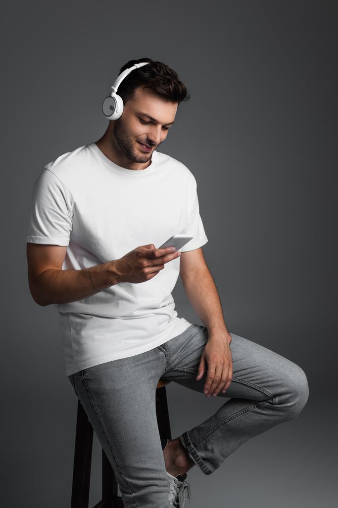 Усміхнений чоловік використовує навушники та смартфон, сидячи на стільці ізольовано на сірому
  - Фото, зображення