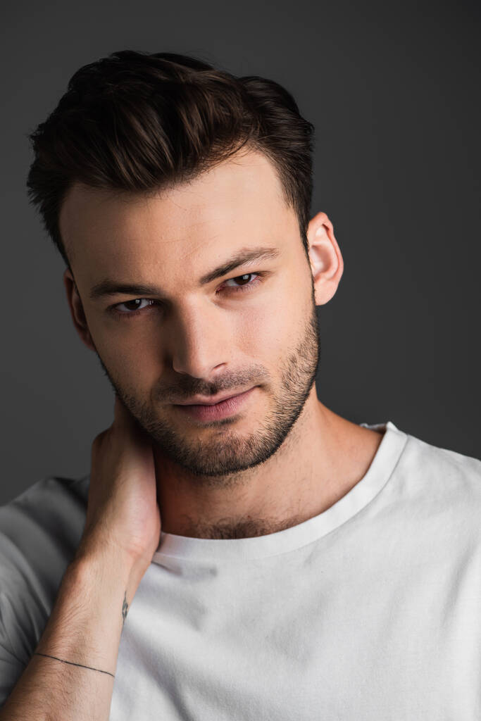 Portrait of young brunette man looking at camera while touching neck isolated on grey  - Photo, image