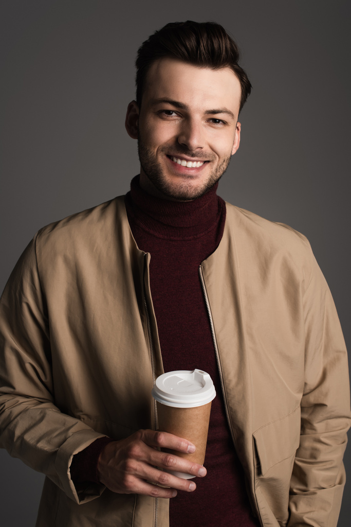 Smiling brunette man in autumn outfit holding paper cup and looking at camera isolated on grey  - 写真・画像