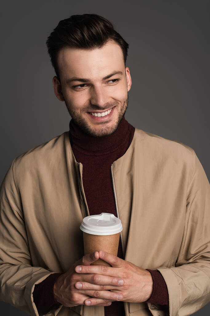 Портрет положительного человека в осенней куртке с напитком на вынос, изолированным на сером  - Фото, изображение