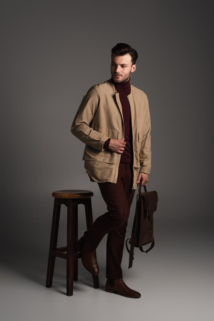 Stylish man in brown autumn jacket holding backpack near wooden chair on grey background - Photo, Image