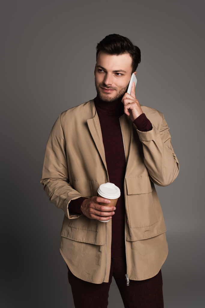 Γενειοφόρος άνδρας με φθινοπωρινή ενδυμασία μιλάει στο κινητό τηλέφωνο και κρατάει καφέ για να απομονωθεί στο γκρι  - Φωτογραφία, εικόνα
