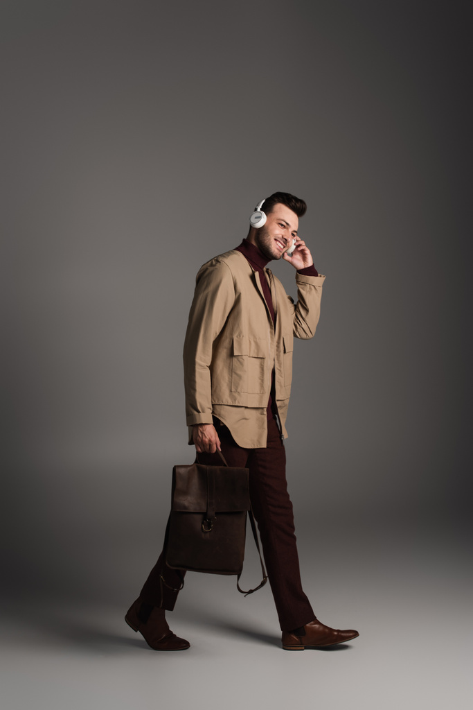 Smiling man in autumn outfit using headphones and holding backpack on grey background - Fotoğraf, Görsel
