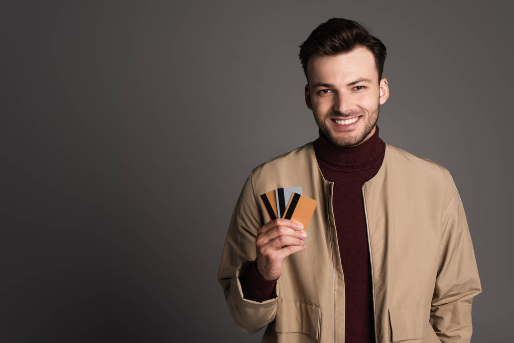 Cheerful man in autumn jacket holding credit cards isolated on grey  - Foto, immagini