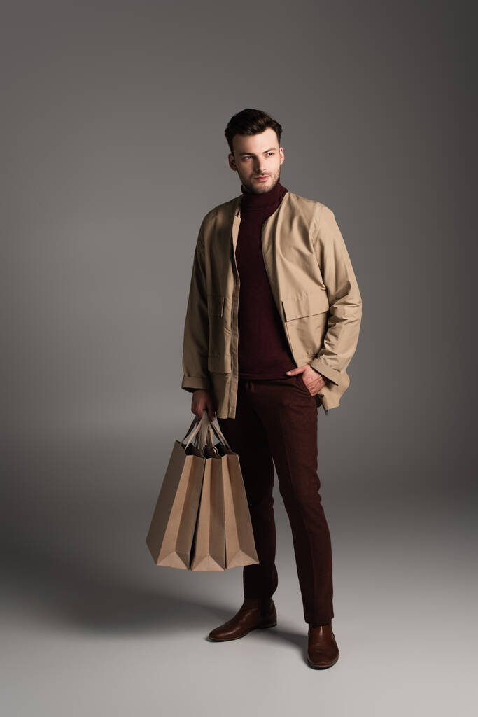 Full length of stylish man in autumn outfit holding shopping bags on grey background - Foto, imagen