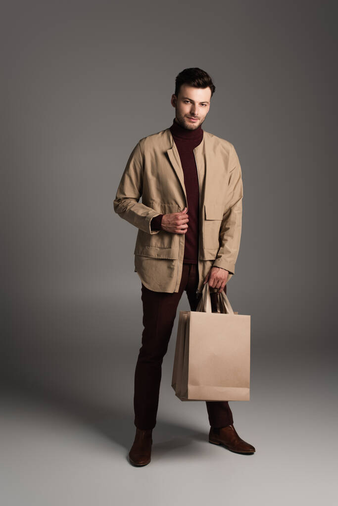 Модный мужчина в коричневой куртке держит сумки на сером фоне - Фото, изображение