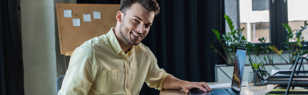 Позитивний бізнесмен в сорочці, використовуючи ноутбук і дивлячись на камеру в офісі, банер
  - Фото, зображення