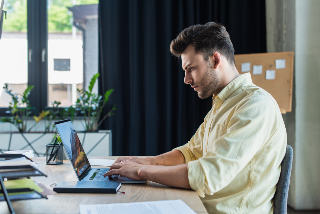 Вид сбоку бизнесмена, использующего ноутбук рядом с документами и ноутбуком в офисе  - Фото, изображение