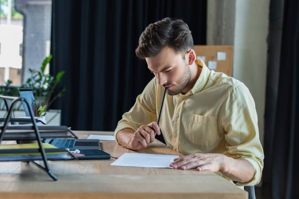 Ευσεβής επιχειρηματίας με πουκάμισο κοιτάζοντας έγγραφο κοντά στο laptop και γραφειοκρατία στο γραφείο  - Φωτογραφία, εικόνα