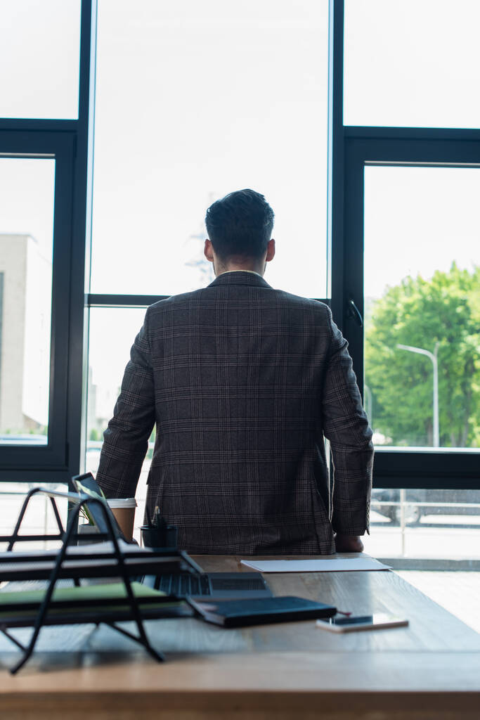 Обратный вид бизнесмена в куртке, стоящего рядом с устройствами и бумагами на рабочем столе в офисе  - Фото, изображение