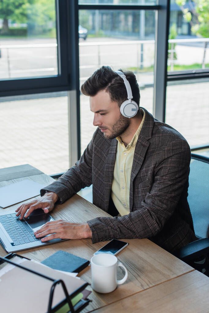 Бізнесмен в навушниках використовує ноутбук біля паперу і чашку на столі в офісі
  - Фото, зображення